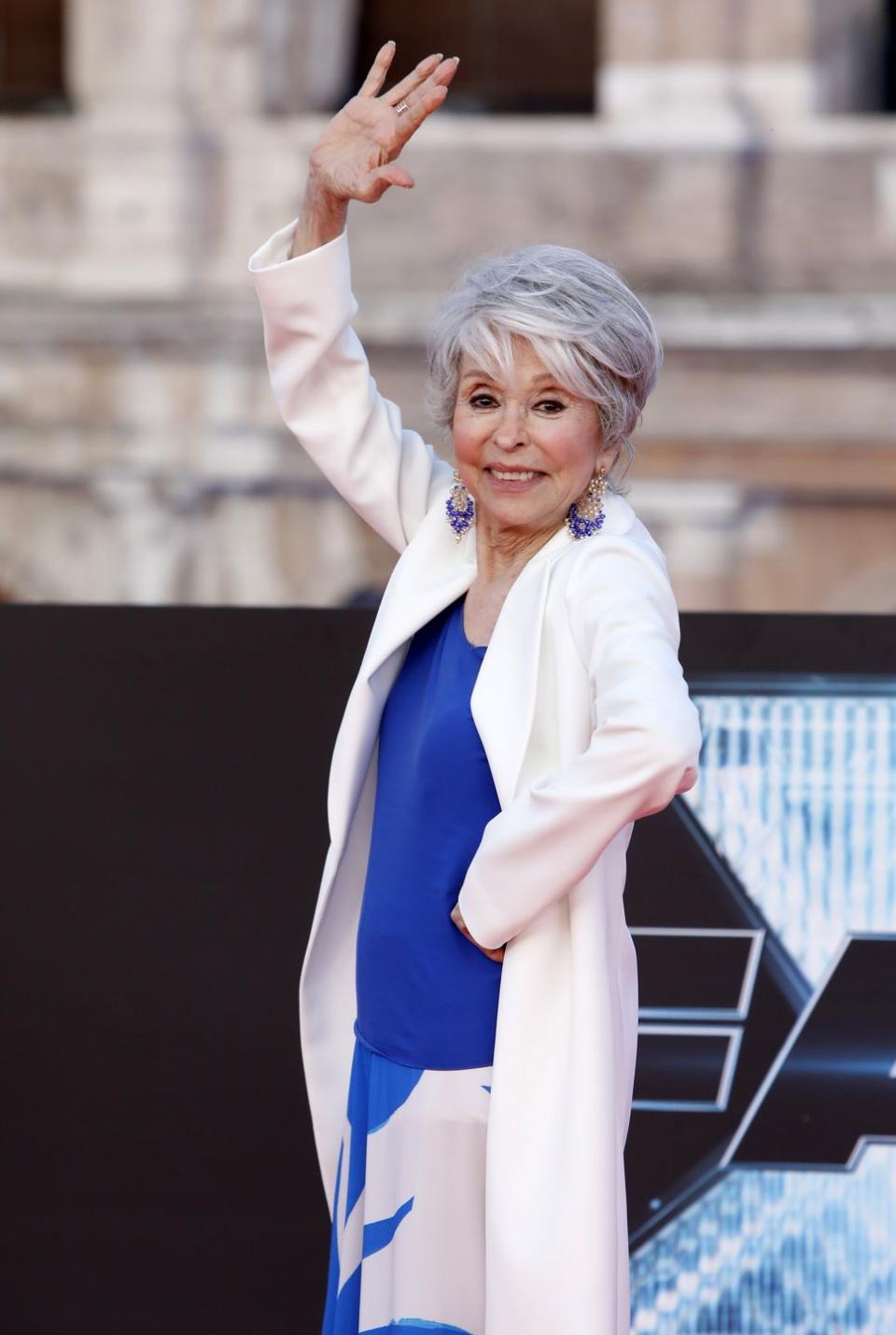 rita moreno smiles and waves at the camera as she looks over her shoulder, one hand is on her hip, she wears a white jacket, blue blouse, and large blue and silver earrings