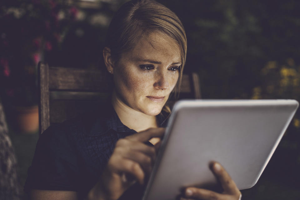 Blondhaired woman using smart device in garden, face is lit by the tablet screen.