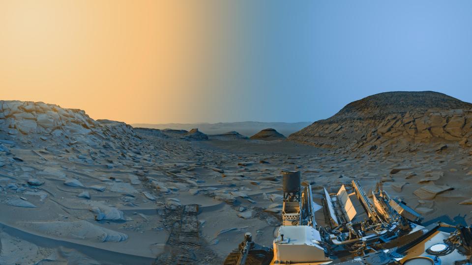 view of a martian valley with a crater rim in the distance and part of the curiosity rover's body in the foreground