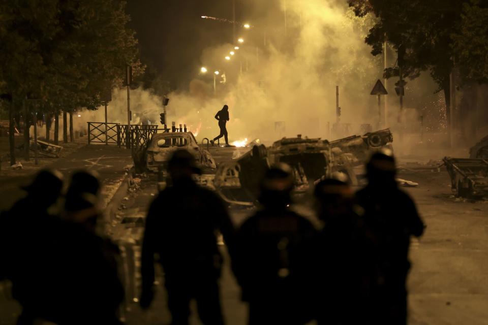 A person runs in the distance as smoke rises. Several people in helmets and dark uniform stand in the foreground