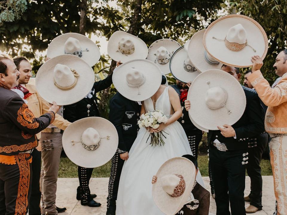 A bride and groom lean together while groomsmen dressed as Charros cover them with sombreros.
