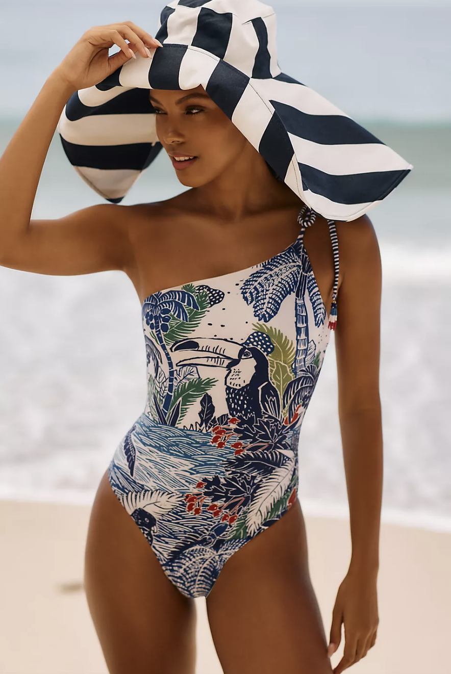 model on beach in striped sun hat and Farm Rio Graphic One-Piece Swimsuit (Photo via Anthropologie)