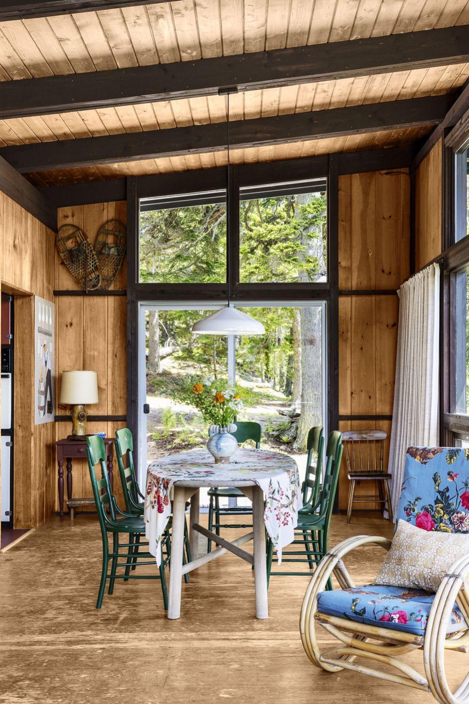 a dining awalls and ceiling are wood paneled with beams, a flowered cloth covered table with painted green chairs, a bentwood rocker with floral cushions, large windows and a sliding glass door