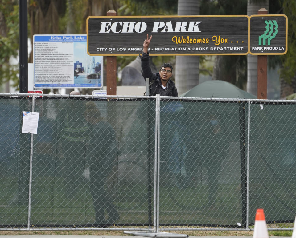 A homeless activist makes a peace sign gesture over a fence, as Los Angeles City sanitation workers move inside the closed perimeter of Echo Park Lake in Los Angeles, Thursday, March 25, 2021. A newly installed fence surrounded the park Thursday after a late-night confrontation between police and vocal demonstrators who oppose the city's effort to remove a large homeless encampment and perform extensive repairs of the site. People who were already in tents at Echo Park Lake were allowed to remain overnight but were given 24-hour notice to leave, Police Chief Michel Moore said in a social media post. (AP Photo/Damian Dovarganes)