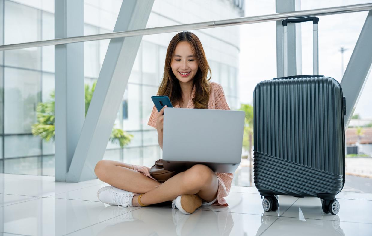 A woman waiting at the airport, working on her phone and laptop, with her suitcase beside her. She's a mobile businesswoman, always on the go. Connectivity is her economy. Air travel concept