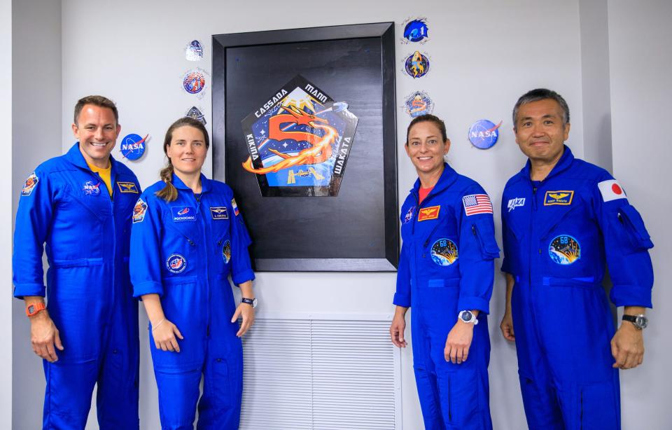 four astronauts in blue jumpsuits post in front of wall of stickers