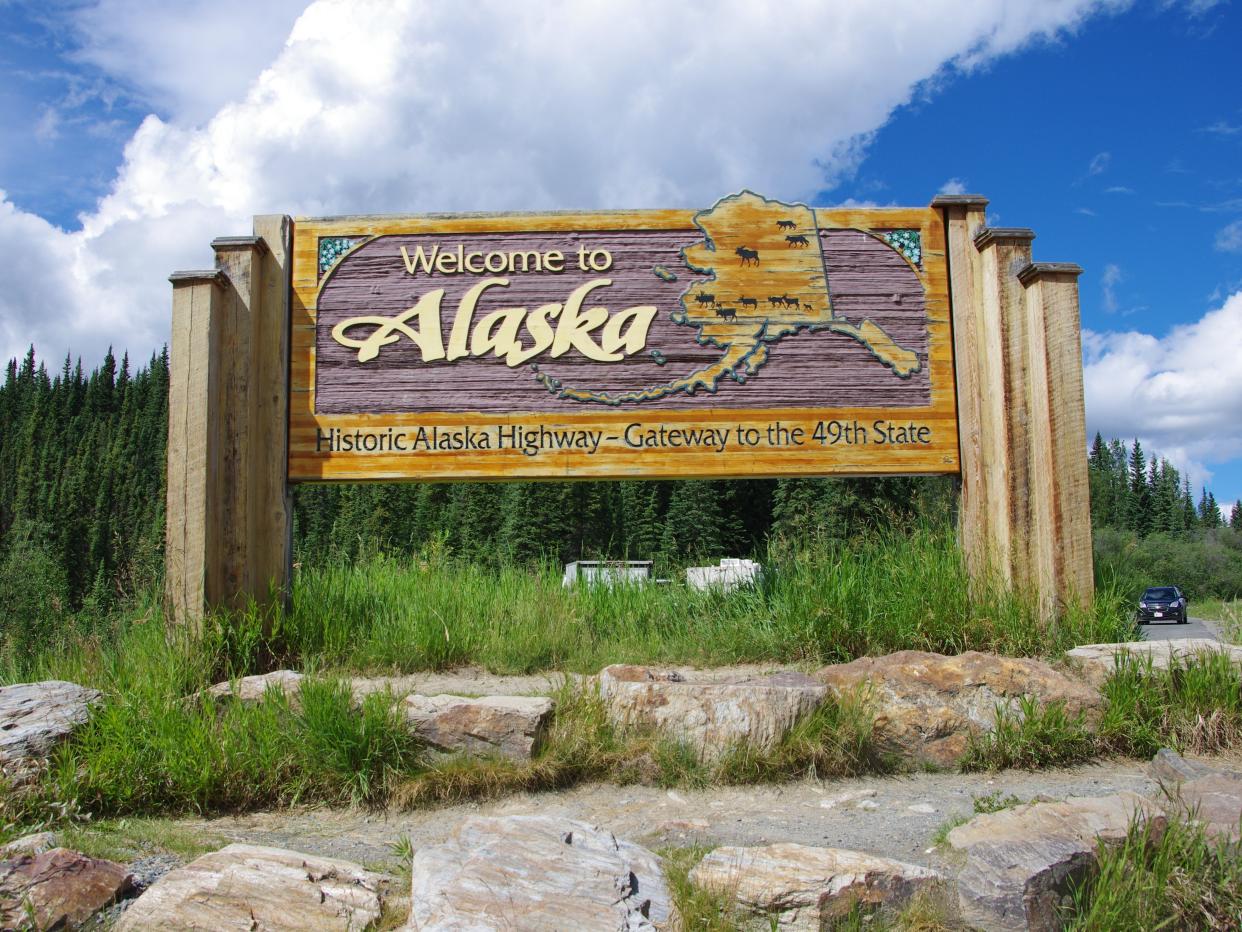 Sign that says "Welcome to Alaska" in front of trees and blue skies