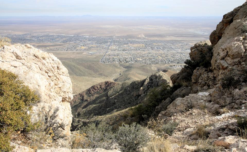 A craggy peak looks onto a rolling range and a large town spread out on the plains below.