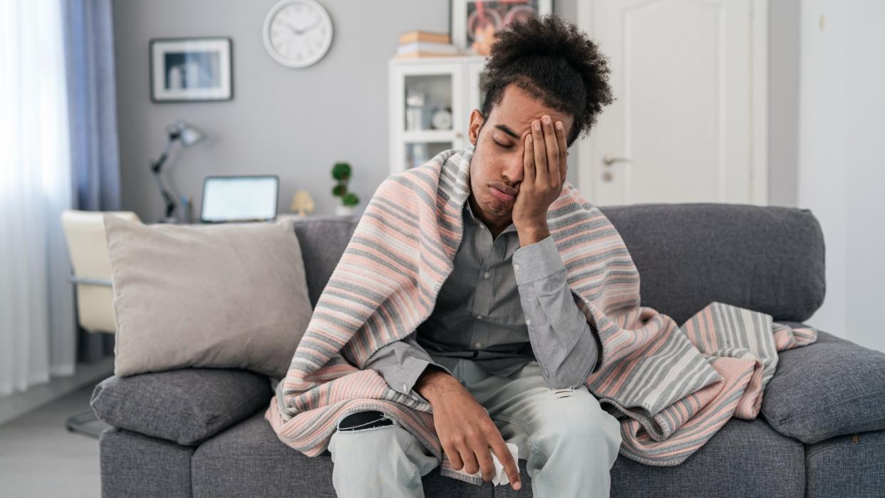  Young african american man, sitting on the sofa at home, having a fever and headache and touching his forehead to check his temperature. 