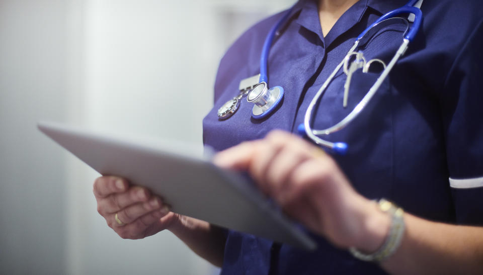 Female surgeon typing on digital tablet in hospital or surgery. She is wearing a dark blue nurse’s top and has her stethoscope around her neck. She is looking at her patients records on her digital tablet and sending emails.