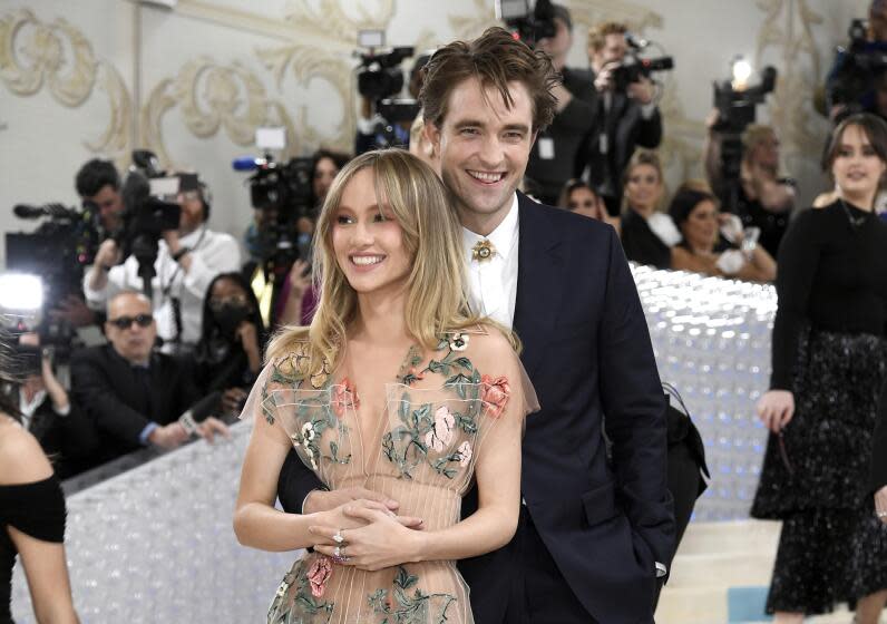 Suki Waterhouse smiles as Robert Pattinson embraces her with one arm, placing his hand along her waist