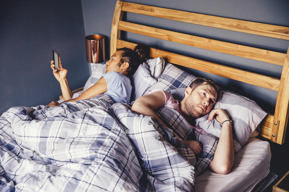 Male couple are lying in bed in the morning. One is lying on his side and is using his smart phone. The other has his arms folded and looks like he is fed up with his partner.