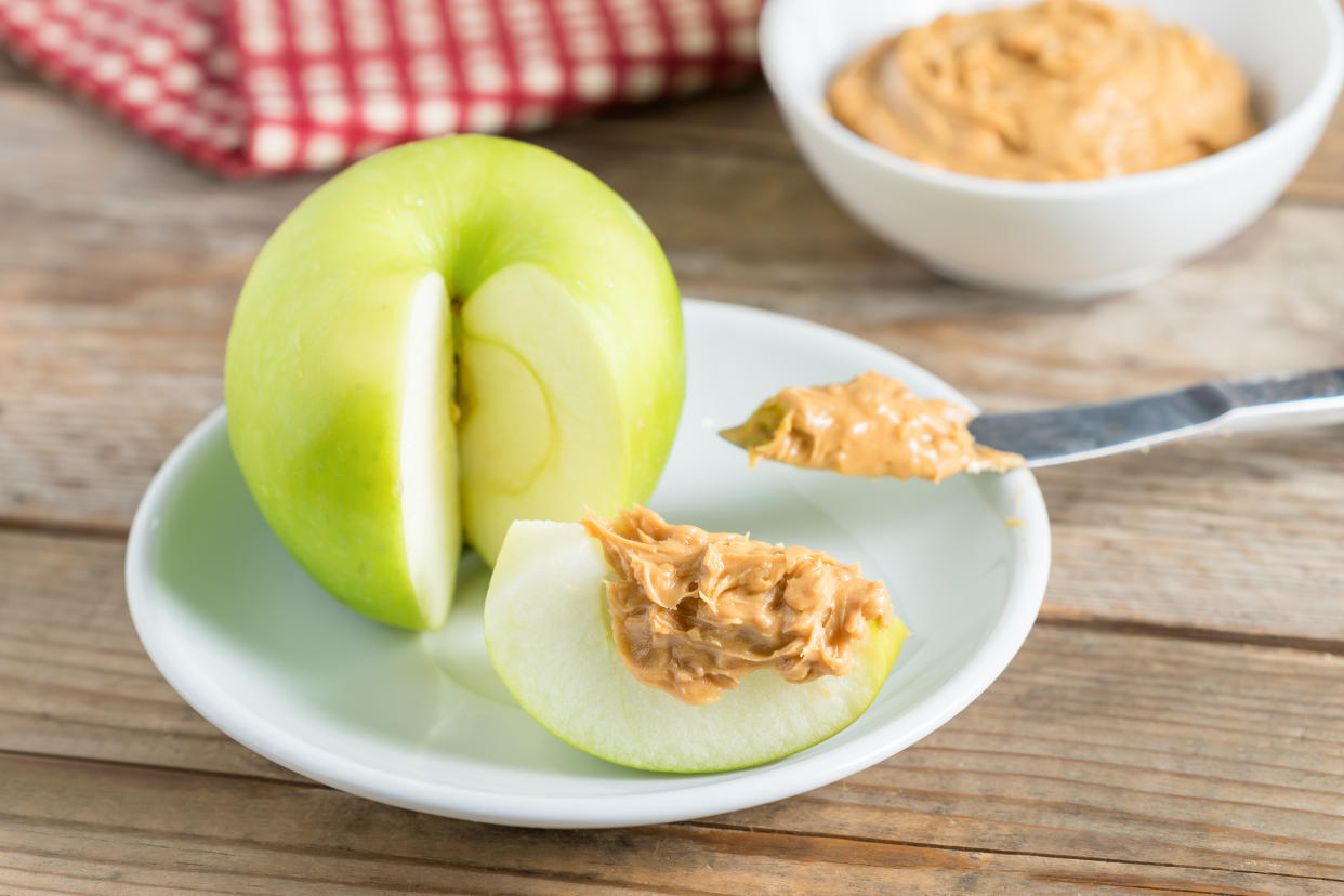 A green apple with a slice removed and nut butter atop it, with a bowlful nearby, sits on a plate with a knife on it.