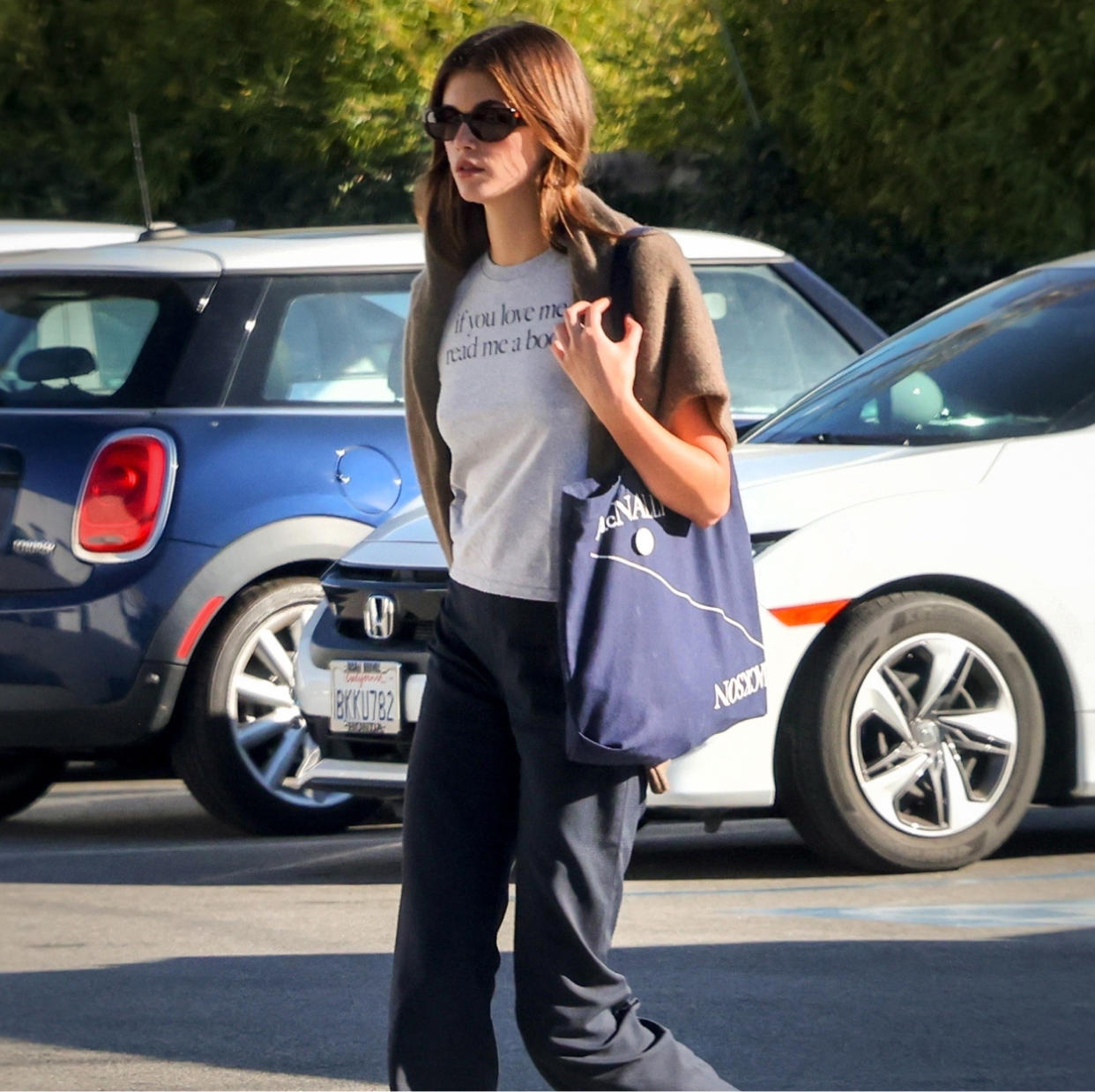  Kaia Gerber wearing a grey tee-shirt, sunglasses, and a blue tote bag. 