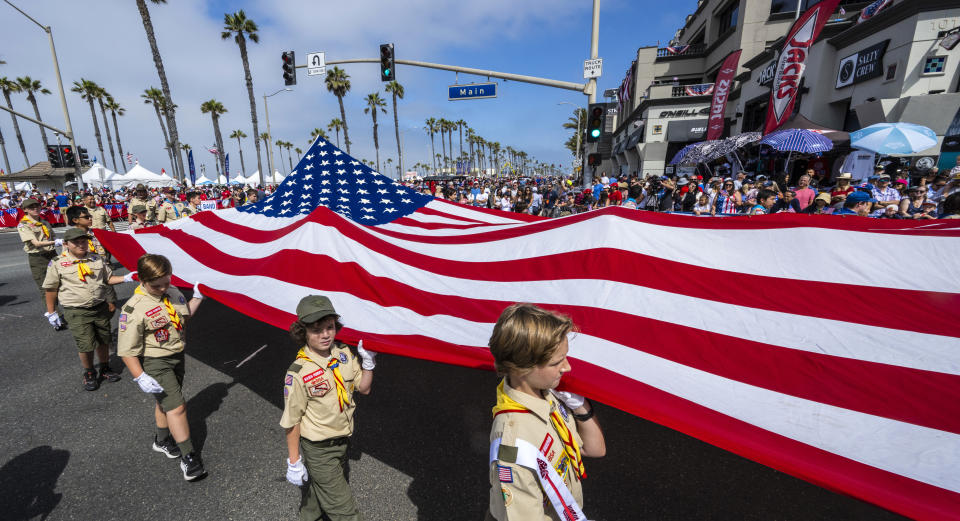 Scouts hold a large American flag during a parade on a palm tree-lined street.