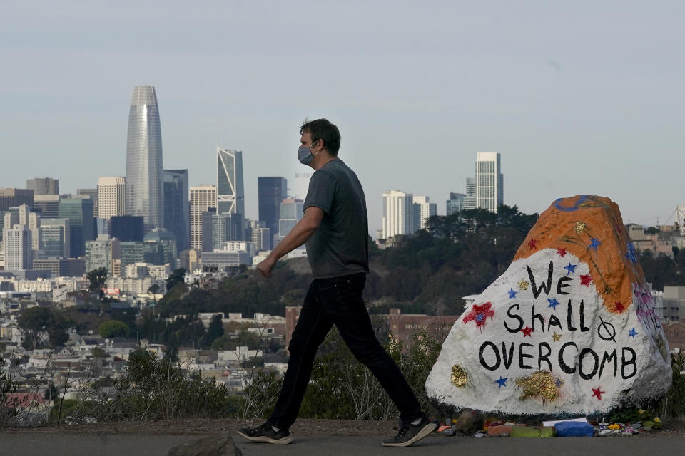 A man wears a mask as he walks past a painted rock on Bernal Heights in front of the skyline during the coronavirus outbreak in San Francisco, Saturday, Nov. 21, 2020. (AP Photo/Jeff Chiu)