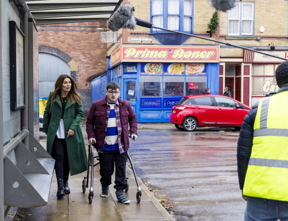 bobby and carla in coronation street, a behind the scenes of filming as a woman and young man with walking aid stroll down the street