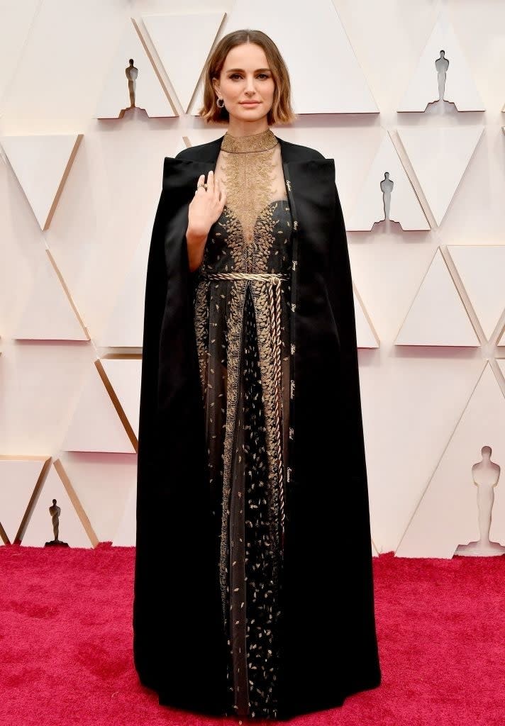 Natalie Portman at an event in a black cape over a gold and black dress, standing on a carpet with statues behind