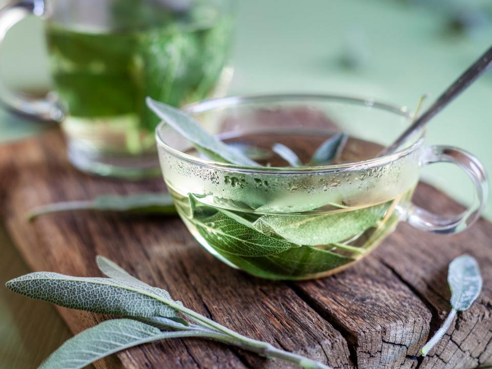 A glass cup of hot water with green leaves in and around it.