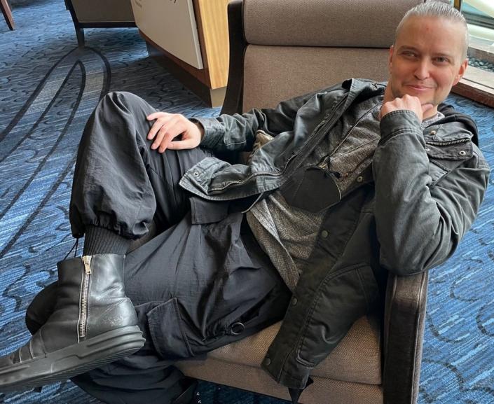 Lucien Greaves smiles while lounging in a chair at the Marriott Copley hotel during SatanCon. He&#39;s wearing a hoodie and a black Barbour jacket.