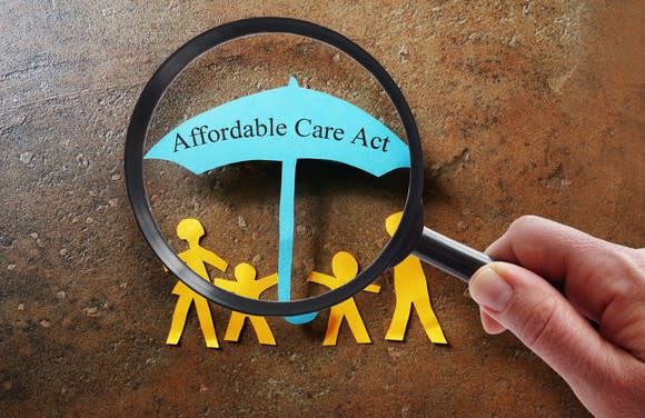 A person holding a magnifying glass looks at paper cut-outs of a family holding an umbrella that reads affordable care act.