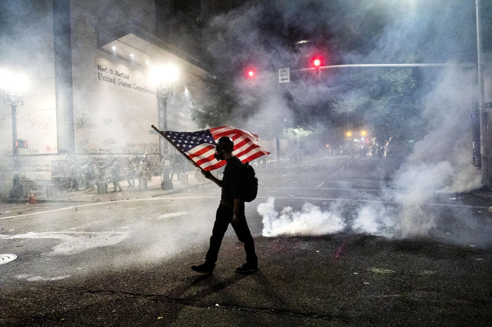 FILE - In this July 21, 2020, file photo, a Black Lives Matter protester carries an American flag as teargas fills the air outside the Mark O. Hatfield United States Courthouse in Portland, Ore. Once hailed as one of the most livable cities in the U.S., Portland, Oregon, is grappling with an uncertain future as it reaches a stunning benchmark: 100 consecutive nights of racial injustice protests marred by vandalism, chaos — and now, the killing of a supporter of President Donald Trump. (AP Photo/Noah Berger, File)