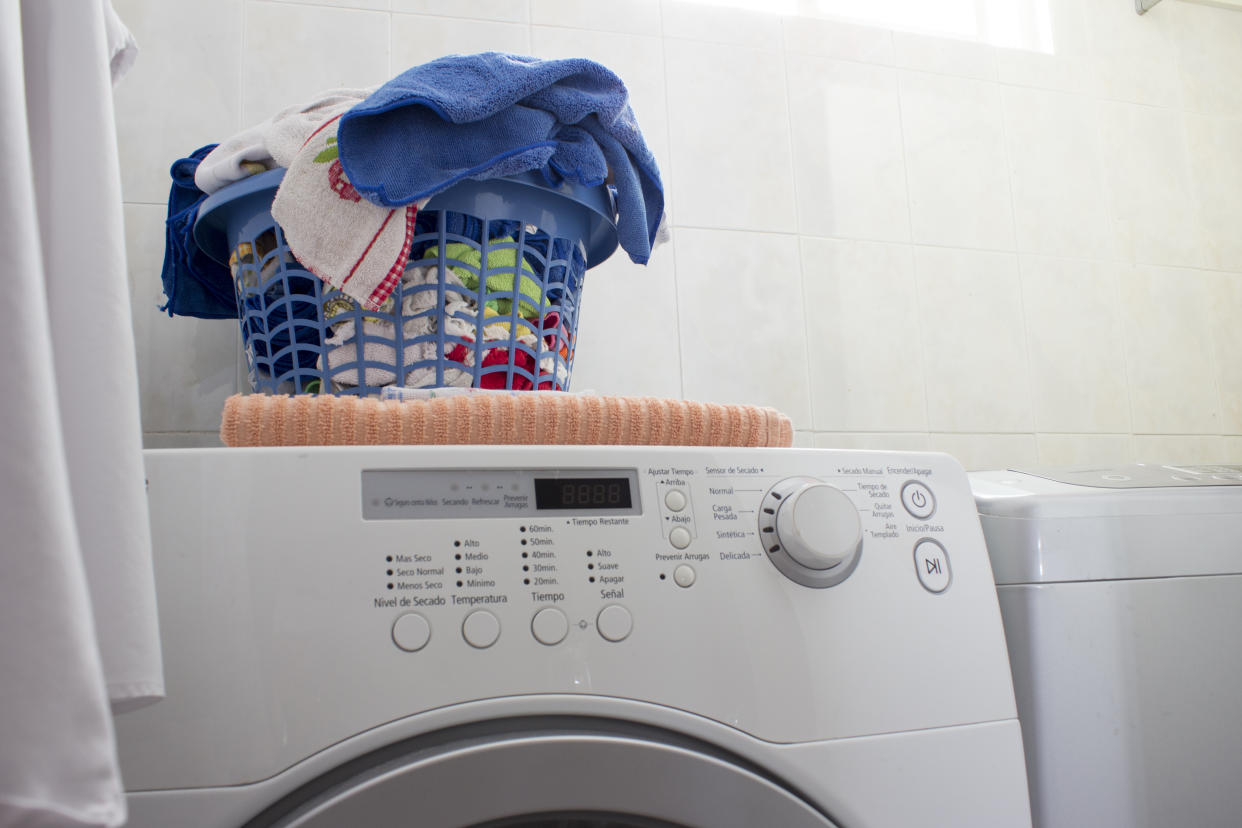 Little laundry area in a domestic house. There is a white shirt hanged at the left. In the center, a clean laundry basket with towels, rags and clothes, over a washing machine.