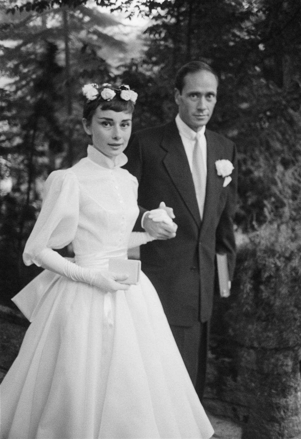 Audrey Hepburn, dressed in a white wedding dress with a crown of flowers, holds hands with her husband, Mel Ferrer.