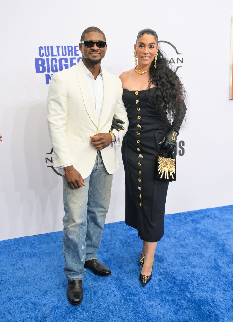 Usher in white blazer and jeans, Jenn Goicoechea in a black, off-the-shoulder dress with gold buttons, standing on a blue carpet at a celebrity event