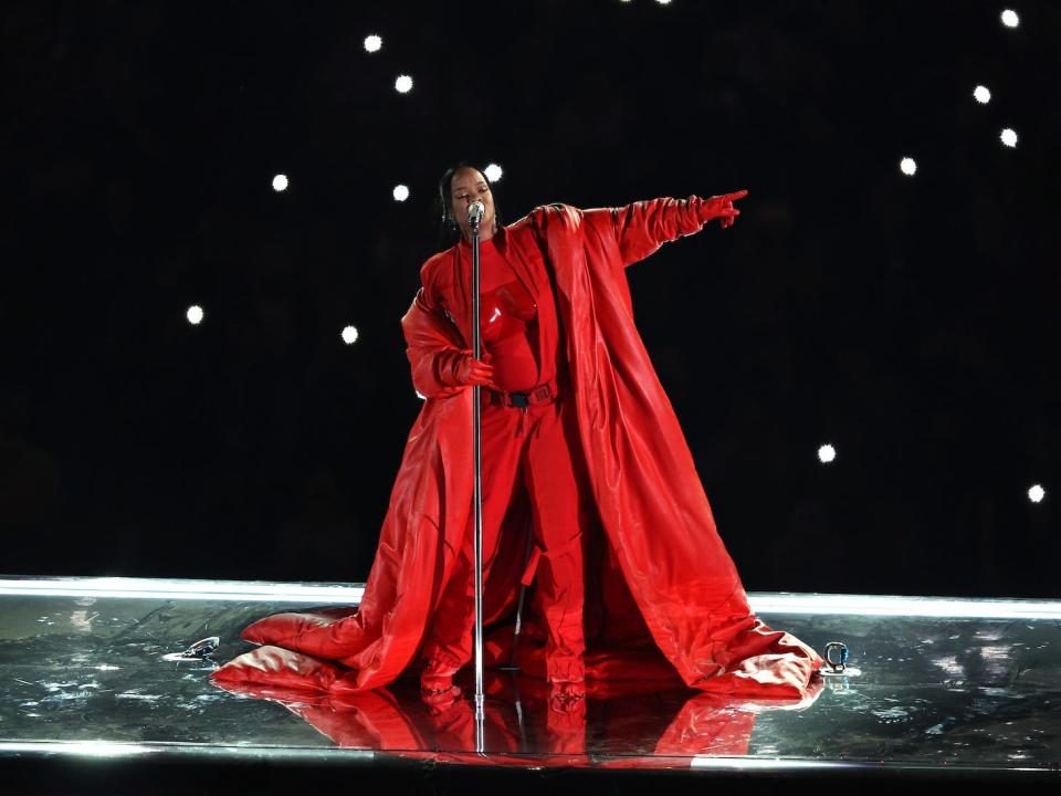 Rihanna sings on a raised platform in a red outfit with glowing lights behind her/