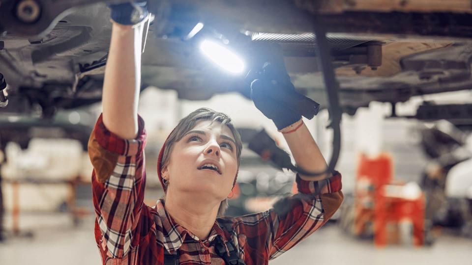 Female technician working with a flashlight under a lifted car.