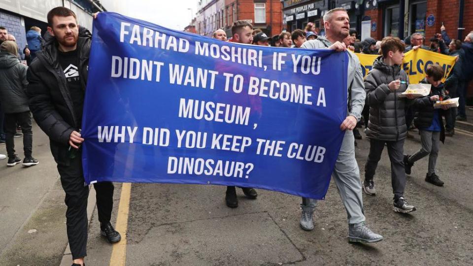 A general view as fans of Everton, holding a banner which reads 'Farhad Moshiri, If You Didnt Want To Become A Museum, Why Did You Keep The Club Dinosaur?'