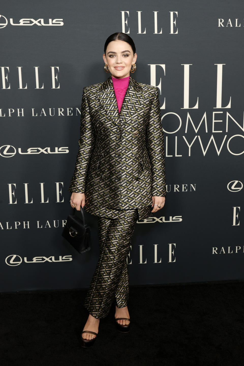 Lucy Hale attends the 27th Annual ELLE Women in Hollywood Celebration at Dolby Terrace. She wears a black suit with a bold golden pattern with a neon pink turtleneck underneath.