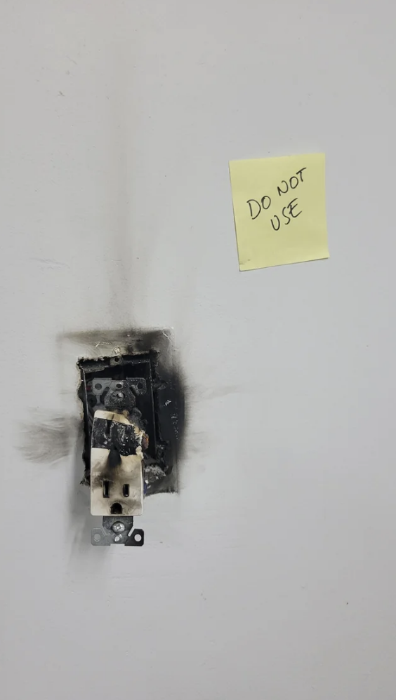 A Post-it Note says "do not use"; it's attached to the wall above an outlet that is falling out of the wall and is covered with burn marks