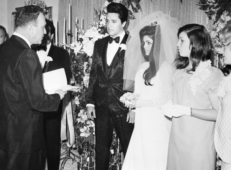 elvis and priscilla presley stand together and face a man who reads from a book he holds, elvis wears a black patterned tuxedo with a white carnation on his lapel, priscilla wears a white wedding dress and veil and holds a small flower bouquet, another woman in a dress stands on the right, a large flower arrangement is in the background