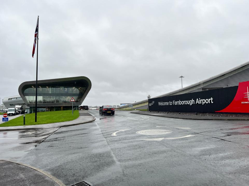 The entrance to Farnborough Airport shows a sign and modern terminal building as a car drives past.