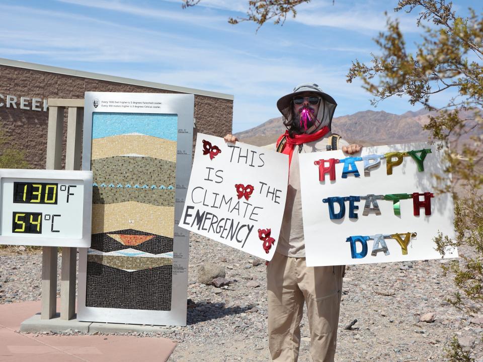A protestor wearing a wide-brimmed hat and a tan suit while holding up two signs that read "This is the climate emergency" and "Happy death day."