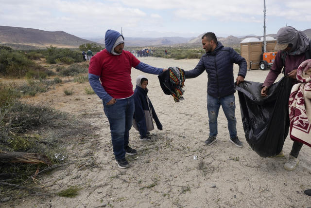 A migrant from Colombia, center, gives a blanket to a father and son, also from Colombia, as the group waits to apply for asylum after crossing the border Wednesday, May 10, 2023, near Jacumba, Calif. The group have been camping just across the border for days, waiting to apply for asylum in the United States. As members of the group get to the front of the line to be escorted into vans, they hand off all warm clothing to those who might still have to camp overnight. (AP Photo/Gregory Bull)