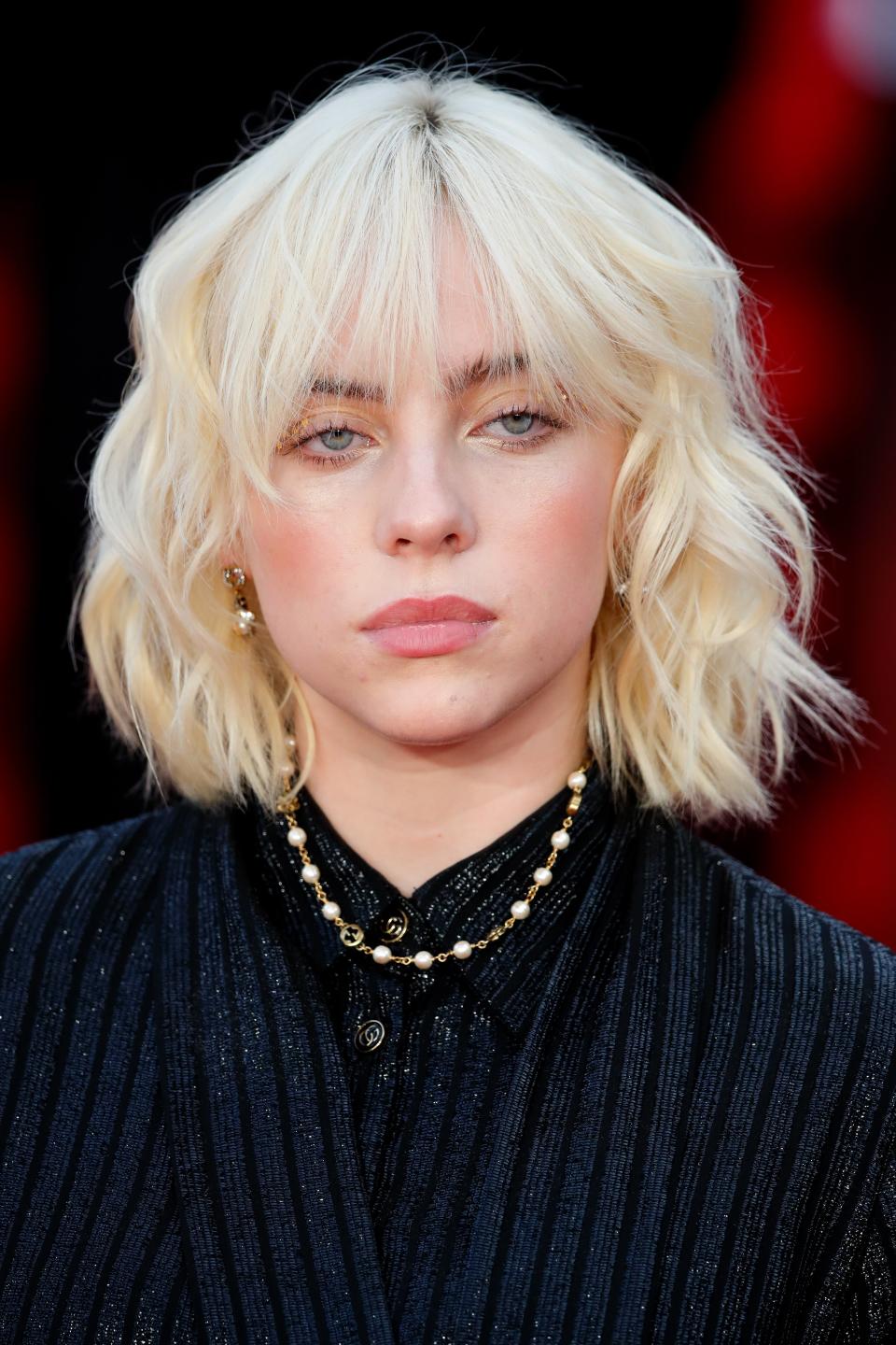 Billie with short blonde hair, wearing a black striped outfit with gold necklace, looking at camera