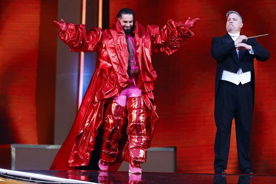 seth rollins strides out onto a stage with his arms raised as he wears a metallic long red jacket and trousers