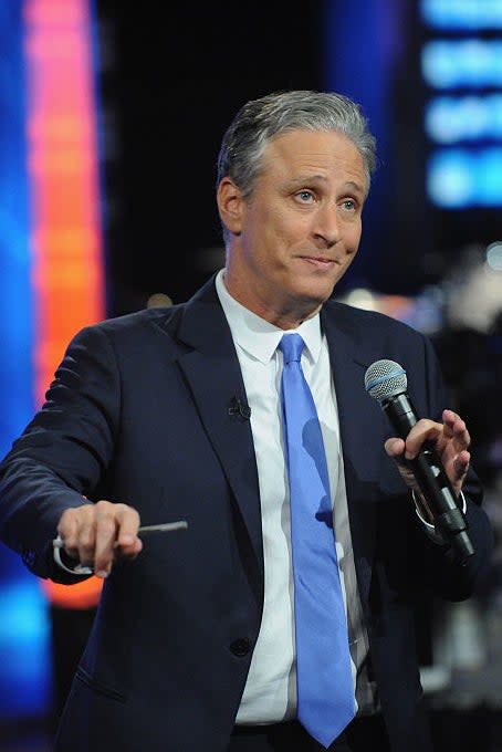 Jon Stewart holding a microphone, dressed in a suit and tie, making a gesture on a stage during a show taping