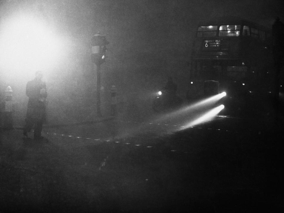 Headlights of a bus faintly glow on Fleet Street in London next to street fog lighting in 1952.