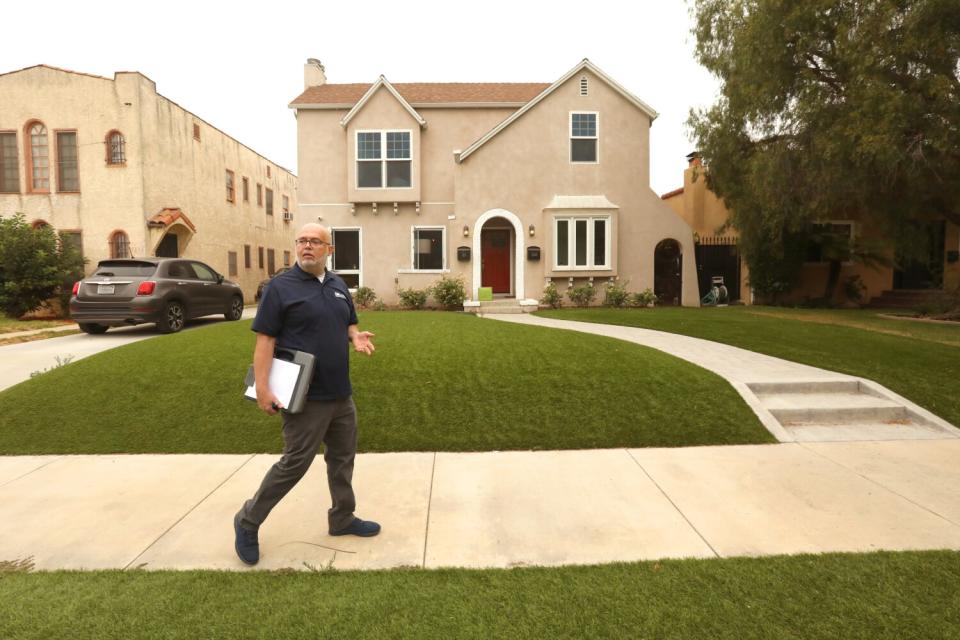 A man in dark clothing gestures while holding a clipboard on the sidewalk outside a home