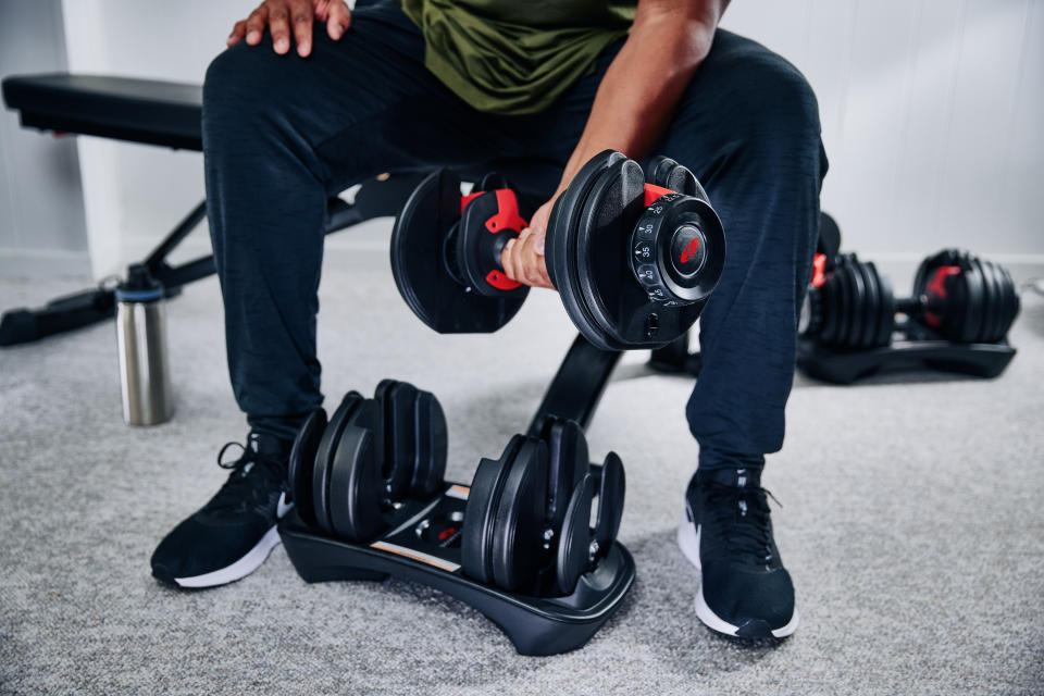 Man, seen from the waist down, doing a bicep curl with a Bowflex SelectTech 552 Adjustable Dumbbell from Canadian Tire while seated on a workout bench in his home gym