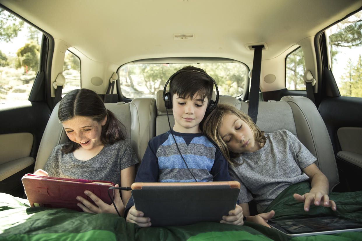 Niños jugando en tableta mientras van en un carro, que parece un road trip. 