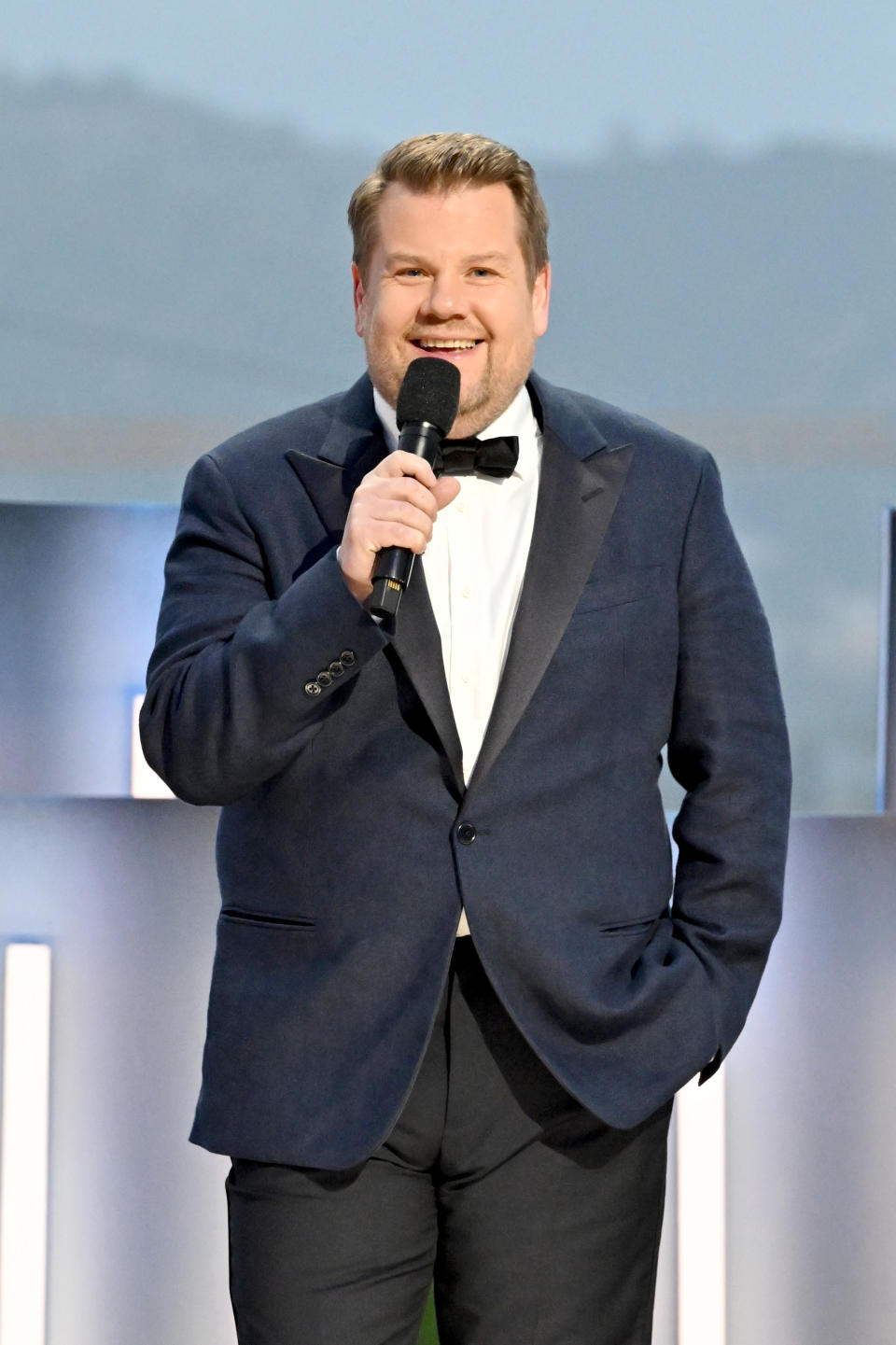 James Corden, in a suit with a bow tie, is holding a microphone and smiling at an event