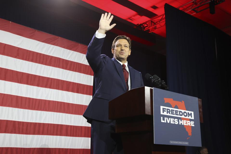 Florida Governor Ron DeSantis, looking jubilant, raises his hand in victory at a podium that says: Freedom Lives Here, over a red map of Florida on a blue background.