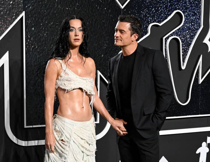 Katy Perry in an asymmetrical fringed top and Orlando Bloom in a black suit hold hands at a red carpet event