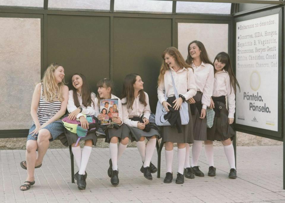 A group of schoolgirls in uniform laugh in a bus stop.