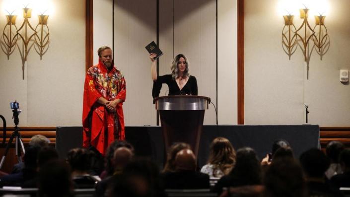 Chalice Blythe, a woman in a long black dress, gestures with a Holy Bible during the opening ceremony for SatanCon. A man in a long red robe stands at her side